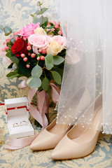 Bridal bouquet of pink, red roses and greenery with satin ribbon, beige women shoes, veil and two golden wedding rings on armchair, copy space. Wedding concept
