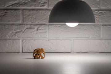 Composition with hand-carved wooden elephant walking through the white table in darkness illuminated by spotlight in front of white brick wall