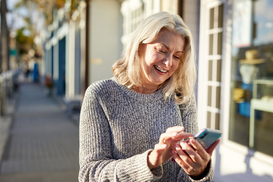 Smiling senior woman using smart phone while standing on sidewalk in city