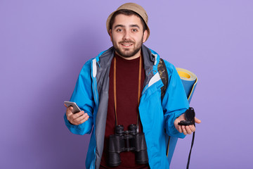 Photo of brave good looking man with beard orientating, choosing better device, holding smartphone and compass, looking directly at camera, having binoculars, travelling alone. Adventures concept.