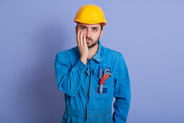 Picture of exhausted tired man rolling his eyes, putting hand on cheeck, wearing yellow helmet and special blue uniform with equipment, standing isolated over lilac background. Tiredness concept