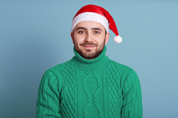 Closeup portrait of pleasant good looking young man standing isolated over blue background in studio, wearing green sweater and red santa claus hat, looking directly. New Year atmosphere concept.