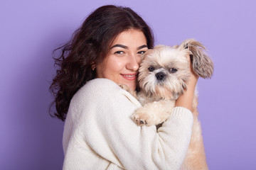 Image of good looking brunette holding Maltese in both hands, standing isolated over lilac background in studio, hugging her favourite pet, being in high spirits, fond of animals. Emotions concept.