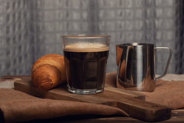 Cup of black americano coffee, french simple croissant and silver color cream sauceboat on wooden board