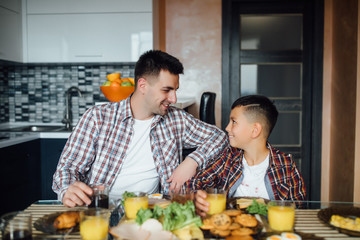 Smiling father and son have a breakfast in kitchen, looking to each other.