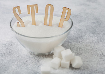 Glass bowl of natural white refined sugar with cubes on light table background with STOp letters. Unhealthy food concept.
