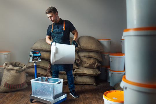 a worker in a uniform pours roasted coffee from a white bucket into a transparent tray on a weighing scale