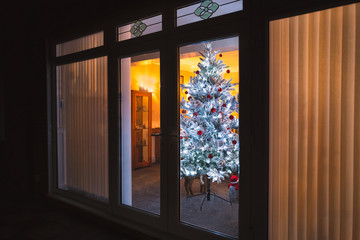 A frosted white artificial christmas tree with red and silver decorations seen through large patio doors with vertical slat blinds and bevel window designs.