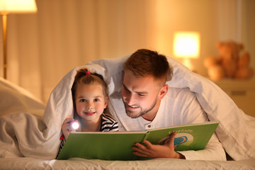 Father and daughter with flashlight reading book under blanket at home