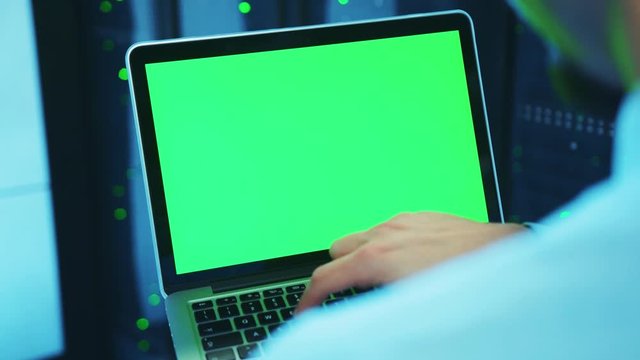 Over the shoulder IT male administrator keyboarding on laptop chrome key green screen, running server diagnostics in database cabinet. Close-up worker with mock-up notebook at data center.