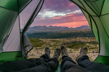 Couple with hiking boots are laying with feet outside the tent opening with a beautiful view over the mountains in the back with amazing sunrise red colored skies. Camping and landscape concept.