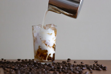 Milk pouring into a glass of iced coffee with beans on a wooden surface