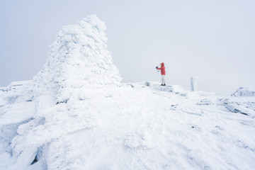 Naklejka na ściany i meble Girl with red hooded jacket and hoodie are taking a selfie next to a frozen cairn on top of a mountain peak covered with snow and fog. Scenery, lifestyle, active and outdoor concept. 