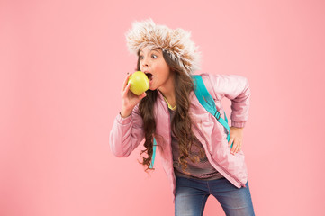 kid pink background. teen daily life. education and happy kids. schoolgirl with books after lesson. school break. small happy girl earflap hat eat apple. end of winter semester. holiday and vacation.