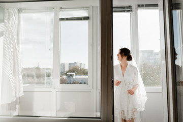Portrait of young gorgeous bride on the balcony. Beautiful girl dresses and gathers for a wedding ceremony in apartments while waiting for the groom. Bride looking at her dress. Bride's morning