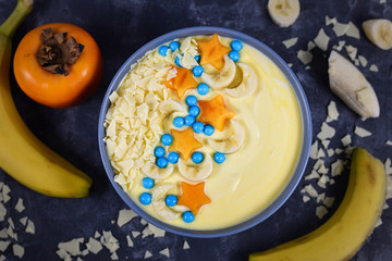 Top view of bowl filled with fruit and yoghurt smoothie decorated with pieces of banana and persimmon  in shape of stars and moons, topped with white chocolate flakes and balls