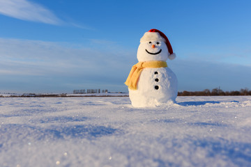 Funny snowman in red santa hat and yellow scalf on snowy field. Christmass and New Year background