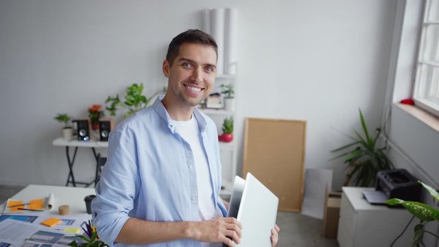 Tilt up of papers and documents on desk and young entrepreneur standing in office, looking at app interface patterns on laptop screen, then closing pc and looking at camera with toothy smile on face
