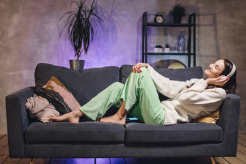 A young woman is lying on the couch in a cozy room listening to music on wireless headphones. Girl...