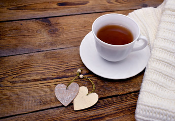 Close up Valentines day composition with white knitted scarf, cup of tea, two decorative heart on wooden desk. Romantic hygge  holiday concept. Copyspace.