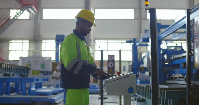 Caucasian bearded engineer uses industrial computer for programming CNC machine on high tech factory. Workman in hardhat working with modern equipment on renovated plant