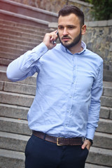 Portrait of a bearded man near the big steps of an old European castle. Businessman talking on the phone. Fashionable guy with stylish clothes.