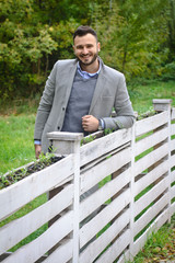 Portrait of a sexy man dressed in a suit. Fashionable guy in the autumn nature near a white wooden fence. Businessman relaxes. Skok photo for design © snb2087
