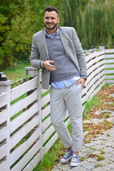 Portrait of a sexy man dressed in a suit. Fashionable guy in the autumn nature near a white wooden fence. Businessman relaxes. Skok photo for design © snb2087