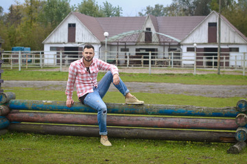 Fototapeta na wymiar Young handsome guy Cowboy. male farmer is sitting on his fence on his ranch. Rural landscapes, countryside. Trees, field, farm. Stock photos for design.
