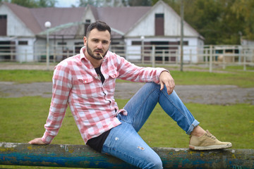 Obraz premium Young handsome guy Cowboy. male farmer is sitting on his fence on his ranch. Rural landscapes, countryside. Trees, field, farm. Stock photos for design.