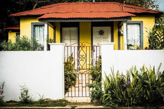 Traditional quaint yellow countryside georgian style house in rural Jamaica with concrete walls, glass louvre windows, zinc roofing, metal gates. Jamaican home with plants in and outside the yard/ lot