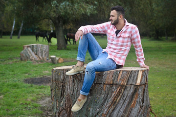 Fototapeta premium Young handsome guy Cowboy. farmer is sitting on a large stump on his ranch. Rural landscapes, countryside. Trees, field, farm. Stock photos for design.