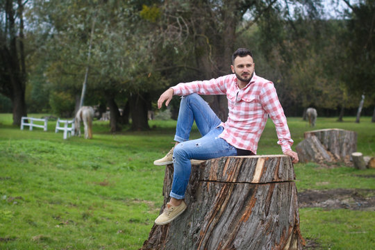 Young handsome guy Cowboy. farmer is sitting on a large stump on his ranch. Rural landscapes, countryside. Trees, field, farm. Stock photos for design.
