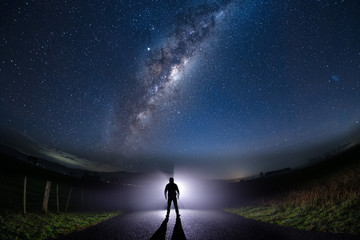 Fototapeta na wymiar A mysterious lost man standing in the middle of the road looking into bright light with milky way starry night sky.