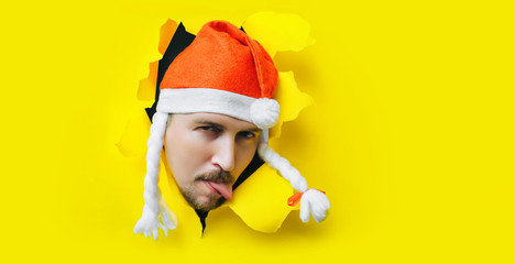 A guy with a beard and a mustache with sticking out tongue in a Santa Claus (Snow Maiden) hat with white pigtails peeps out of a torn hole in yellow paper. ?oncept of Christmas, New Year. Copy space.