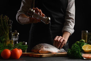 Chef cooks fresh fish on the background with ingredients, salts it before cooking, freezing in motion, advertising photo for seafood sale, cookbook for recipes