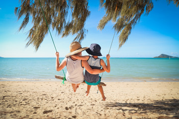 Kids swinging together on log swing at the beach in tropical north Queensland.  Slow living on tropical family holiday in paradise with sand and sea. - Powered by Adobe
