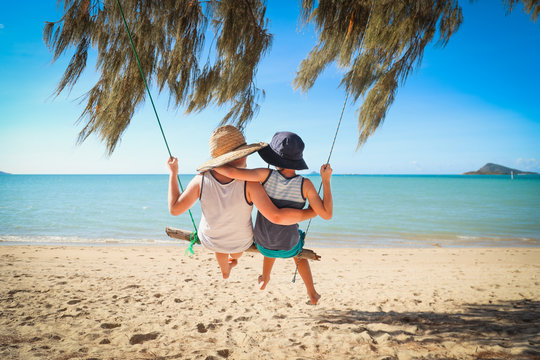 Kids swinging together on log swing at the beach in tropical north Queensland.  Slow living on tropical family holiday in paradise with sand and sea.