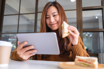 An asian woman holding and eating whole wheat sandwich while using tablet pc