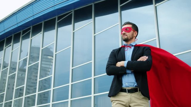 Portrait of handsome young businessman in suit wearing superman costume outdoors standing alone near office building, red cape is wavering in the wind.