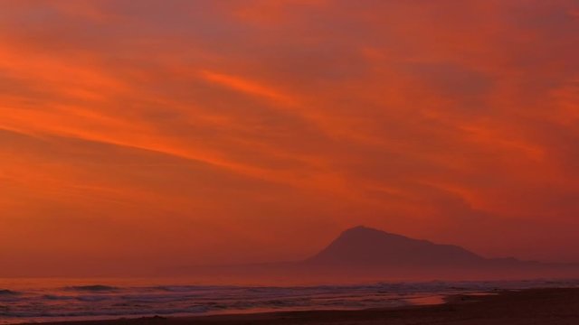 Dramatic coastal seascape of orange dawn skies over sandy beach and mountain, zoom