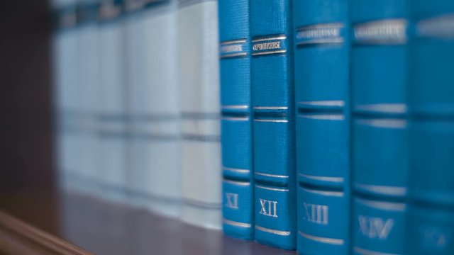 Collection of volumes of books of blue color by different writers on a wooden bookshelf. Closeup. Dolly shot