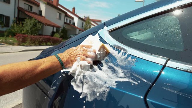 SLOW MOTION, CLOSE UP, DOF: Unrecognizable person runs a soapy sponge along the side of blue car. Person slaps the shiny blue car with a soap water filled squeegee. White foam splashes over car.