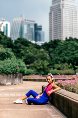Fototapeta na wymiar Full-length portrait of fit blonde in stylish bright sportswear sitting in city park after training. Fitness woman listening to music via headphones while relaxing. Sporty girl concept. Vertical shot