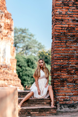 Fototapeta premium Portrait of attractive tourist girl on vacation looking at camera while having photoshoot in white dress near old ruins. Summer trip, tourism, adventures concept. Vertical shot