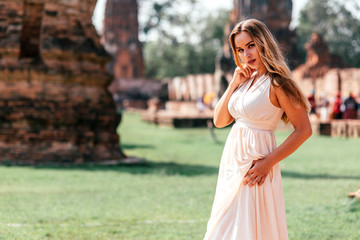 Portrait of attractive tourist girl on vacation looking at camera, admiring asian landscape while having photoshoot in white dress near old ruins. Summer trip, adventures concept. Horizontal shot