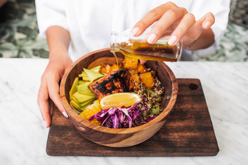 Woman pouring olive oil in meal with fried salmon fish steak, quinoa, avocado, corn, cabbage salad and baked pumpkin in wooden bowl. Healthy organic food concept. White marble table surface. - 308627503