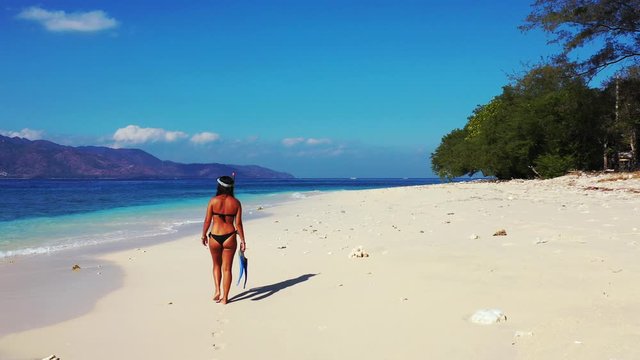 Attractive woman in the mini bikini with snorkeling mask on the head and fins in the hand walking along the sandy beach, Bahamas