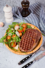Grilled beef steak with fresh arugula, tomato and Parmesan salad on a plate on a grey concrete background. Vertical  photo.