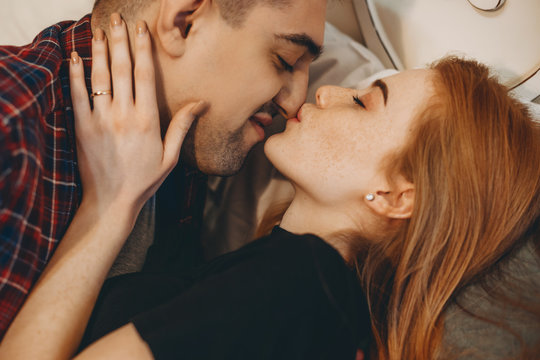 Close up of a charming young female with red hair and freckles kiss her boyfriends face while embracing on a bed at home closely.
